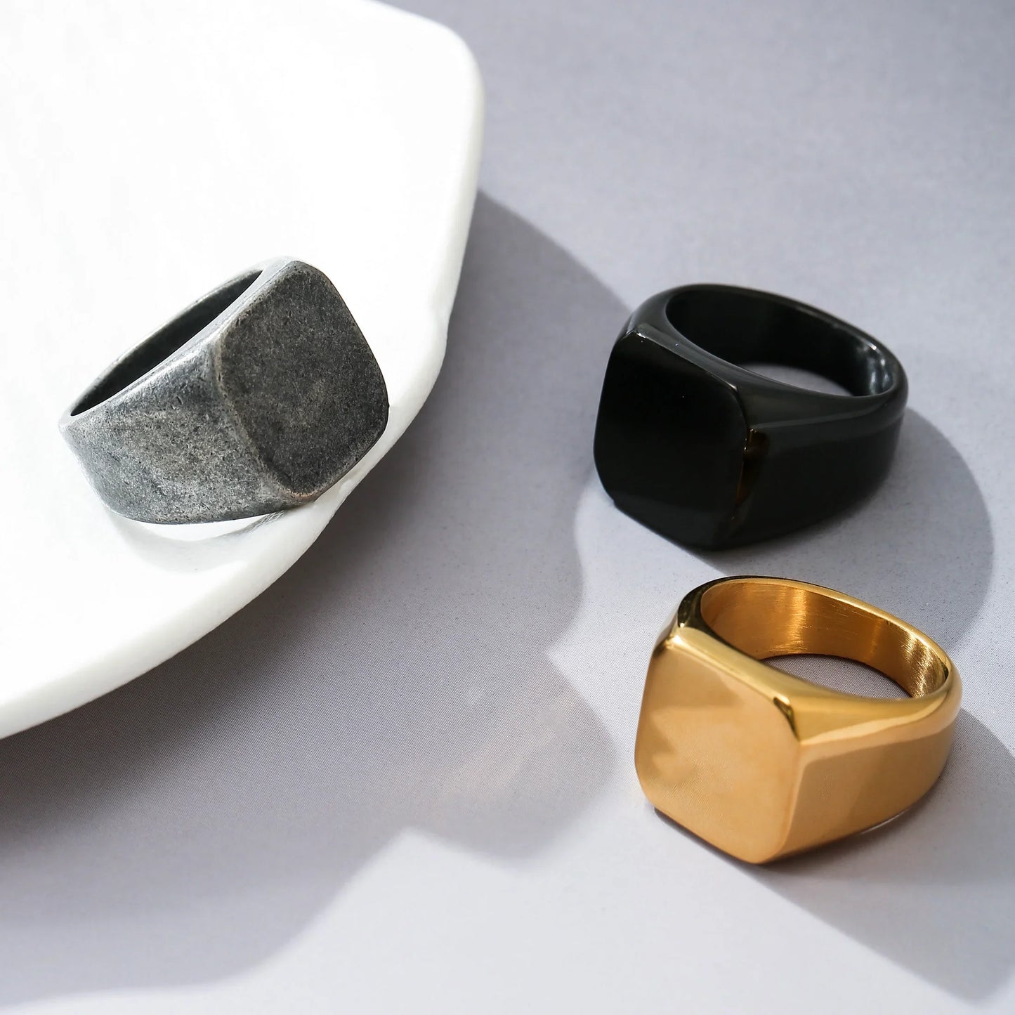 Three minimalist signet rings in gold, matte black, and brushed silver on a gray surface.