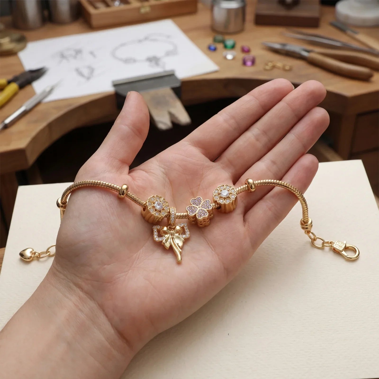 Hand holding gold charm bracelet with clover and bow diamond details, jewelry workshop background