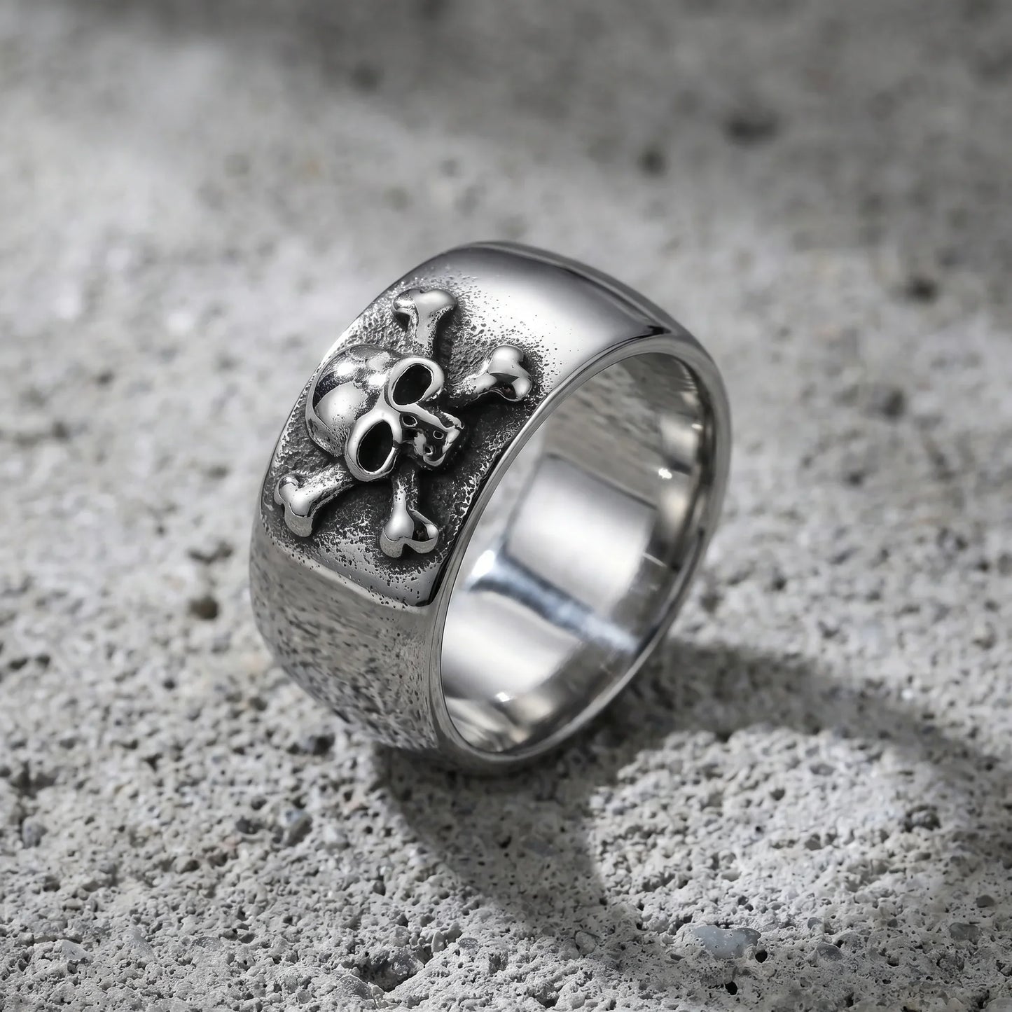 Wide silver ring with 3D skull and crossbones design on textured concrete surface