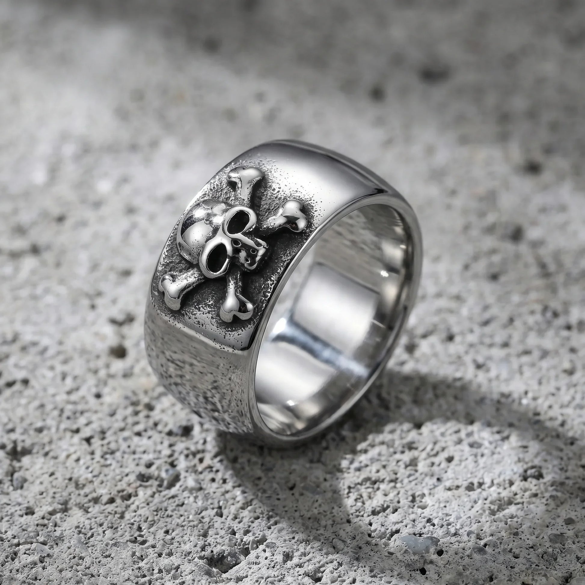 Wide silver ring with 3D skull and crossbones design on textured concrete surface