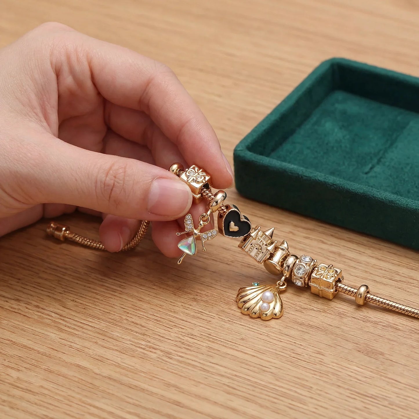 Hand holding gold charm bracelet with fairy, heart, castle, and seashell charms on wood table