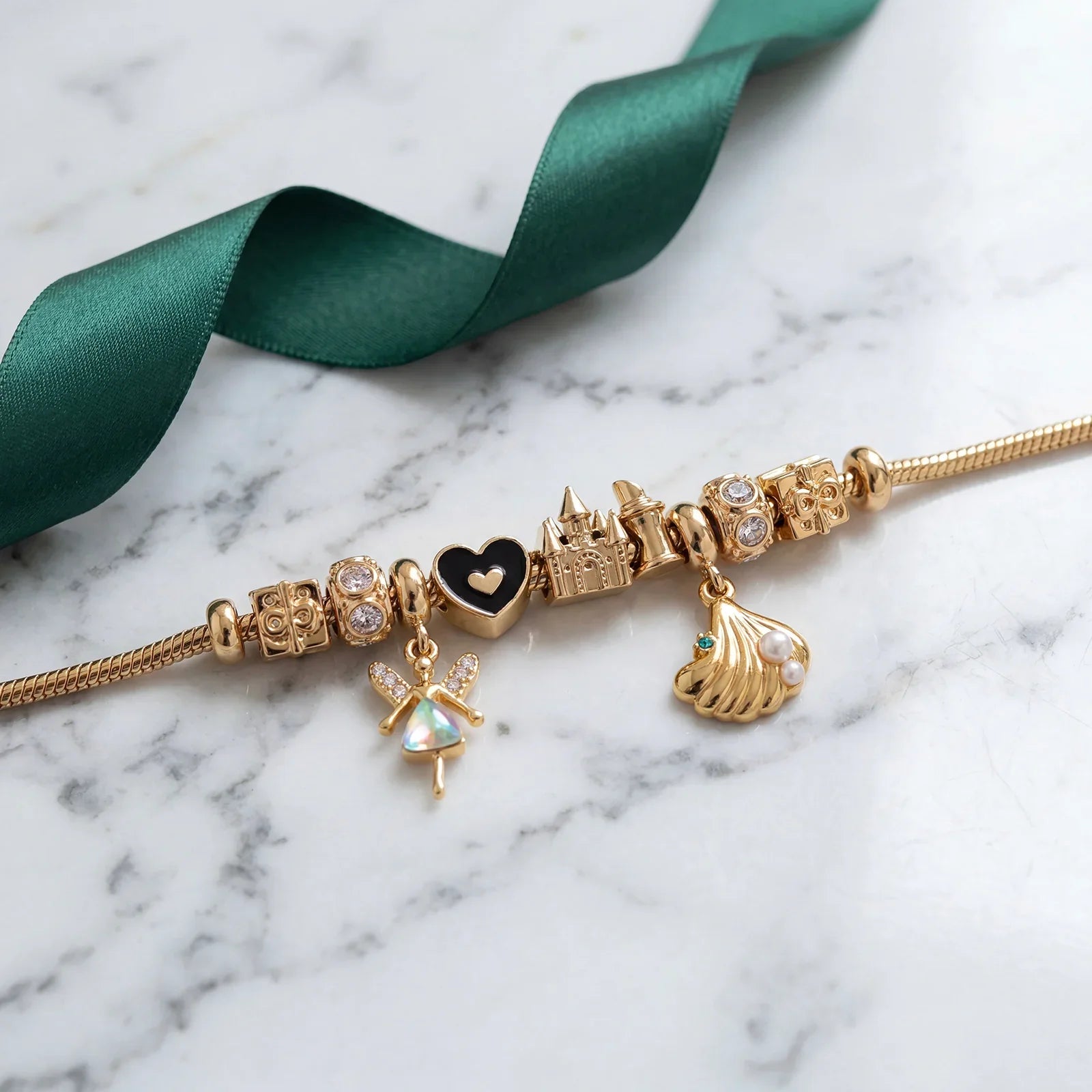 Gold charm bracelet with castle, heart, fairy, and seashell charms on marble surface