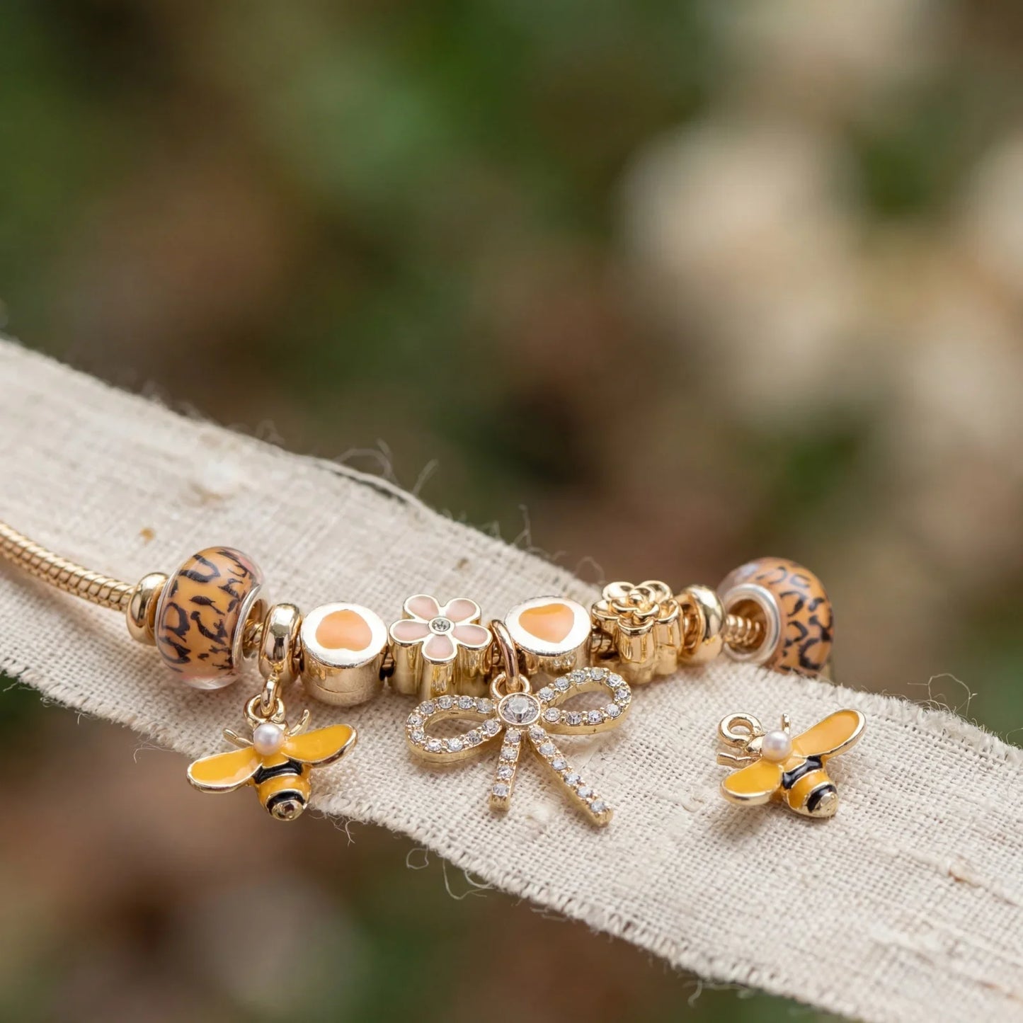 Gold charm bracelet with bee, bow, and flower charms on a neutral fabric background
